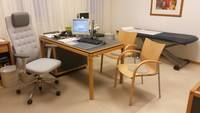 in der Mitte des Raumes befindet sich ein Arbeitstisch mit zwei Stühlen. Dahinter befindet sich eine Behandlungsliege.