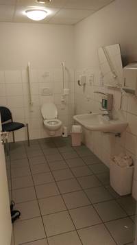 Auf dem Foto ist die Toilette zu sehen. Links von der Toilette befindet sich ein Mülleimer. Rechts von der Toilette befindet sich ein Stuhl. Außerdem befindet sich das Waschbecken von der Toilette aus auf der linken Seite. 